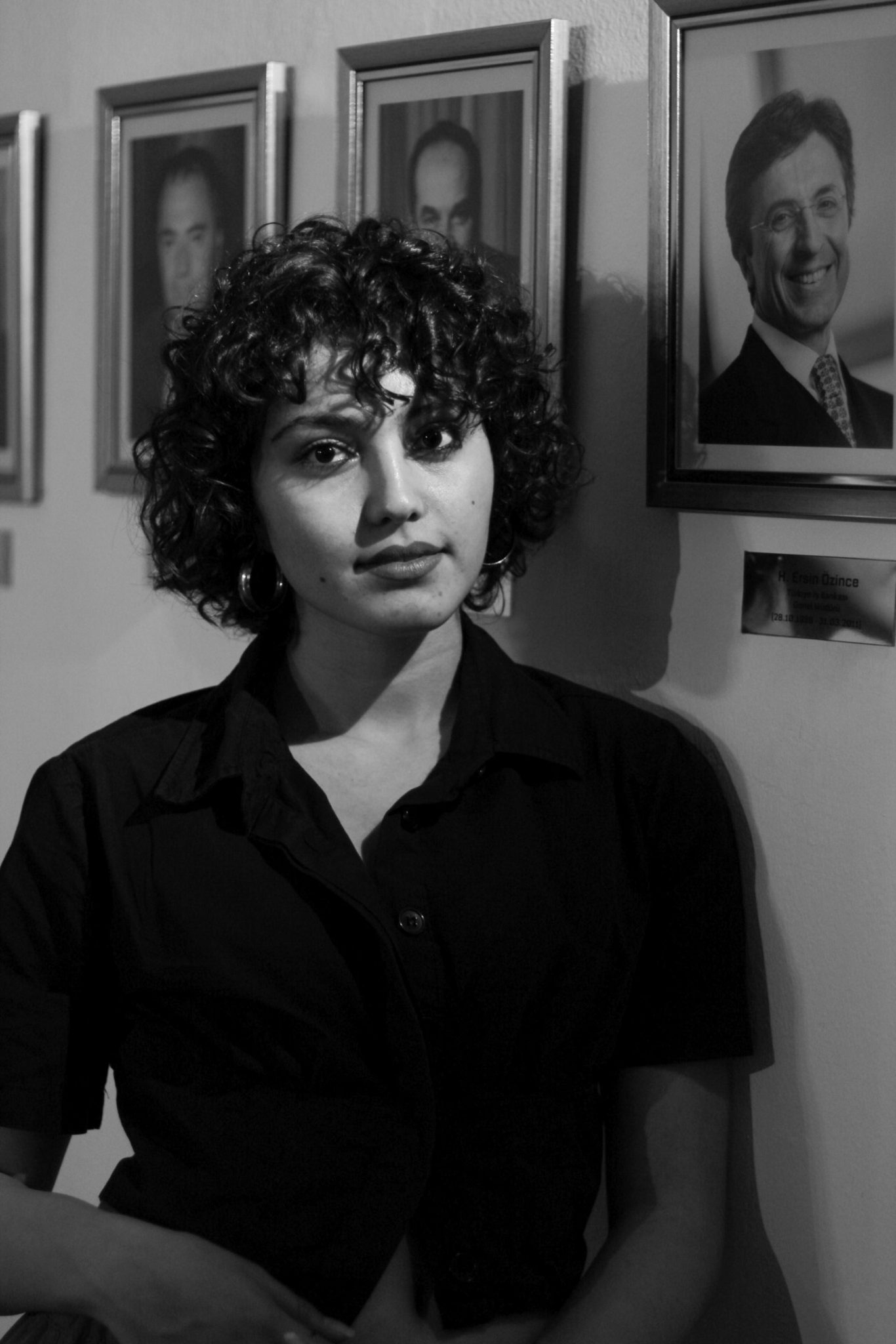 Black and white portrait of a woman with curly hair standing against a wall with framed portraits.